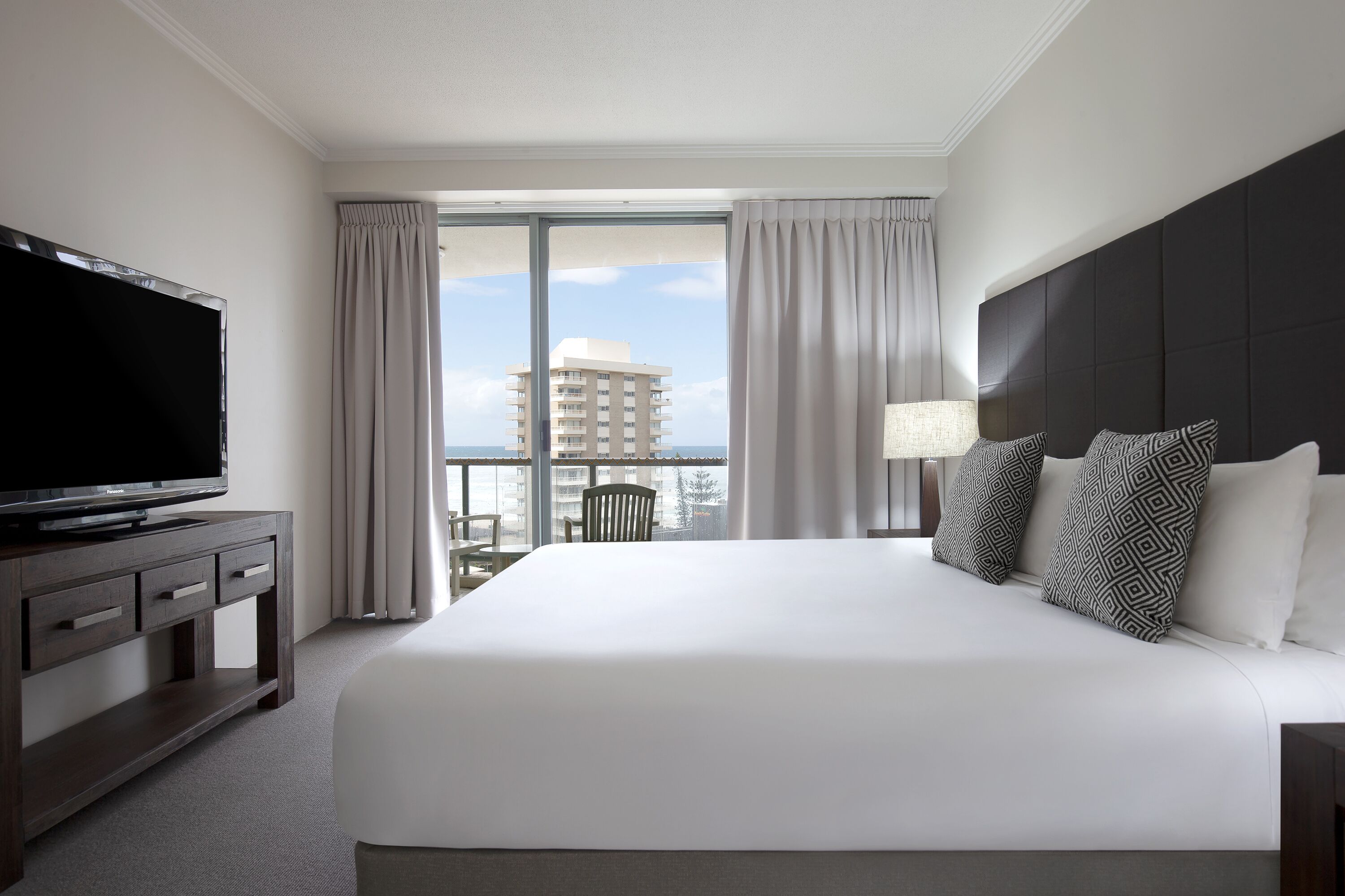 Habitación de hotel con una cama grande, un televisor de pantalla plana sobre una consola de madera, un balcón con cortinas y vistas a un edificio.