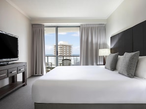 Habitación de hotel con una cama grande, un televisor sobre un mueble de madera, un balcón con cortinas y vistas a un edificio.