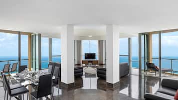 A modern living room with a large TV, a dining area with a glass table, and a balcony with a view of the ocean.