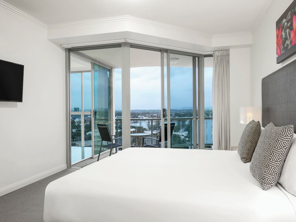 Habitación de hotel con una cama grande, vistas de la ciudad a través de ventanales del suelo al techo y un balcón con mesa y sillas.