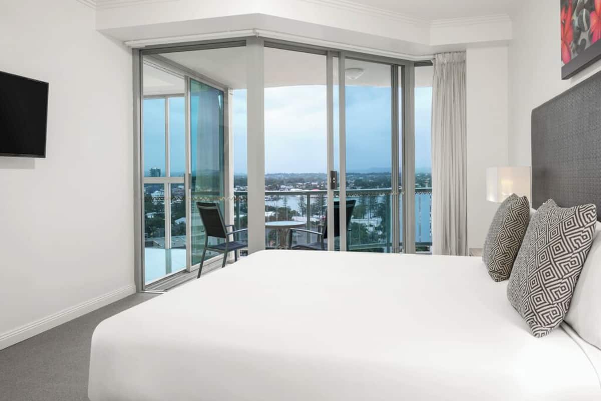 Habitación de hotel con una cama grande, vistas de la ciudad a través de ventanales del suelo al techo y un balcón con mesa y sillas.