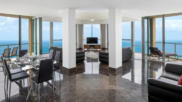 A modern living room with a large TV, a dining area with a glass table, and a balcony with a view of the ocean. - Mantra Sun City Surfers Paradise (Gold Coast)