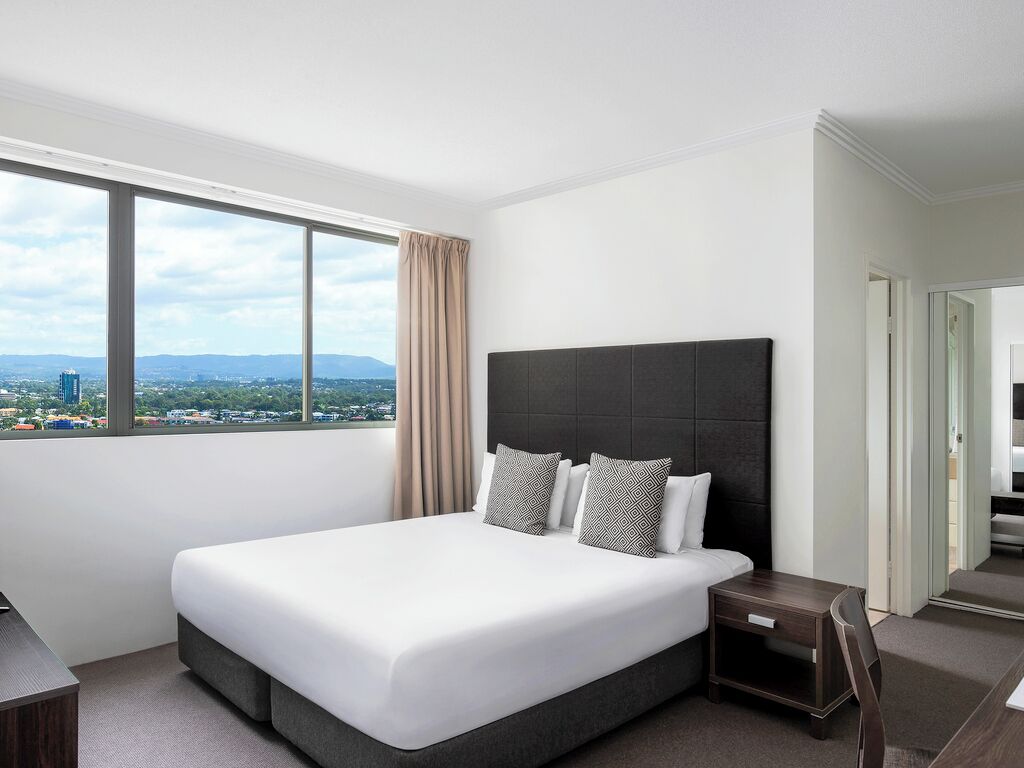 Habitación de hotel moderna con una cama grande, una mesita de noche y vistas a la ciudad a través de un amplio ventanal.