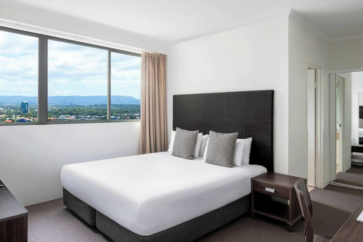 Moderni hotellihuone, jossa on suuri sänky, yöpöytä ja suuri ikkuna, josta avautuu näkymä kaupunkiin.
