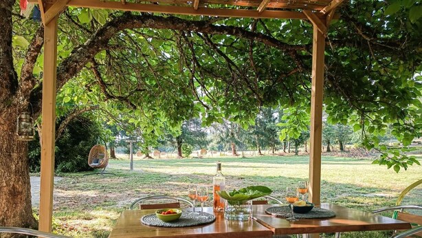 En udendørs spiseplads med pergola, borde, stole og udsigt til en grøn park.