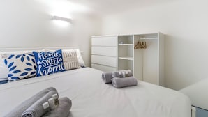 A neatly made bed with rolled towels and toiletries, a white wardrobe with hanging space, and a bedside table with a lamp.