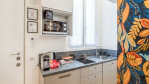 A compact kitchen with a microwave, coffee maker, and sink. There are framed certificates on the wall and a window with curtains.