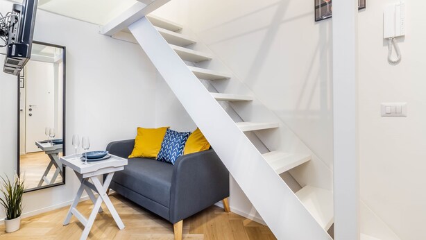A modern living room with a grey sofa, yellow and blue patterned cushions, a small table with glasses, and a white staircase.
