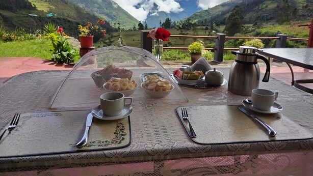 Una mesa dispuesta con un calentador de té, tazas y una cesta de galletas, con vistas a un paisaje montañoso.