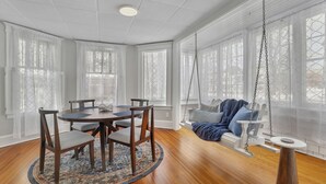 Una habitación con una mesa redonda de madera, seis sillas, un columpio y ventanas grandes con cortinas translúcidas.
