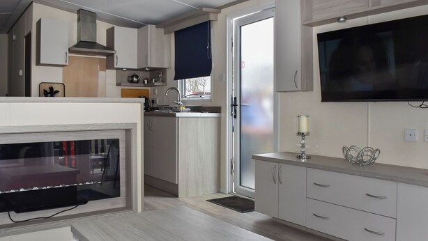 A modern kitchen with a built-in oven, a dining area with a table, and a TV mounted on the wall. - This lovely caravan is perfect for couples, friends, or families. (Sandown)