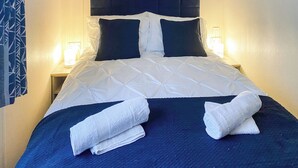 A neatly made bed with a dark headboard, white bedding, and two bedside lamps. - This lovely caravan is perfect for couples, friends, or families. (Sandown)