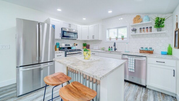 Una cocina moderna con armarios blancos, electrodomésticos de acero inoxidable y una isla de mármol con dos taburetes de madera.