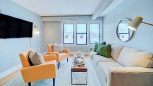 A living room with a flat-screen TV, two orange armchairs, a beige sofa, and a coffee table with books. - Lovely Mid-Century Modern - Apt B (Muskegon)