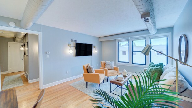 A modern living room with a sofa, armchairs, and a coffee table. There is a TV mounted on the wall, a lamp, and a large window providing natural light. - Lovely Mid-Century Modern - Apt B (Muskegon)