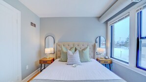 A bedroom with a large bed, two bedside tables with lamps, a mirror, and a window with a view of a snowy landscape. - Lovely Mid-Century Modern - Apt B (Muskegon)