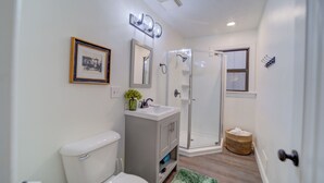 A bathroom with a toilet, sink, mirror, shower, and a framed picture on the wall. - Parisian Botanical - Apt D (Muskegon)