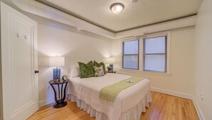 A bedroom with a bed, bedside tables, a nightstand, a window with blinds, and a door. - Parisian Botanical - Apt D (Muskegon)