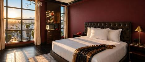 A large bed with a dark headboard, a nightstand with a lamp, a dresser with a mirror, and a view of the city through large windows.
