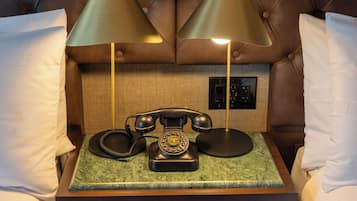Une chambre d’hôtel avec une tête de lit capitonnée, un téléphone à cadran d’époque posé sur une table en marbre vert, et deux lampes de chaque côté.