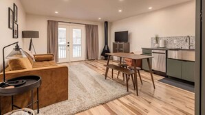 A modern living room with a sofa, a coffee table, and a dining area with a table and chairs. There is a kitchen area with a stove, a sink, and cabinets. - Slopeside Suite at Alyeska - Ski In Ski Out (Girdwood)
