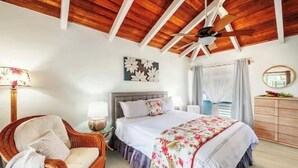 A bedroom with a wooden ceiling, a bed with floral bedding, a wicker chair, and a bedside table with a lamp. - Chic Aloha Studio in Superb Princeville, w/AC & WiFi 
Explore Beautiful Napali! (Princeville)