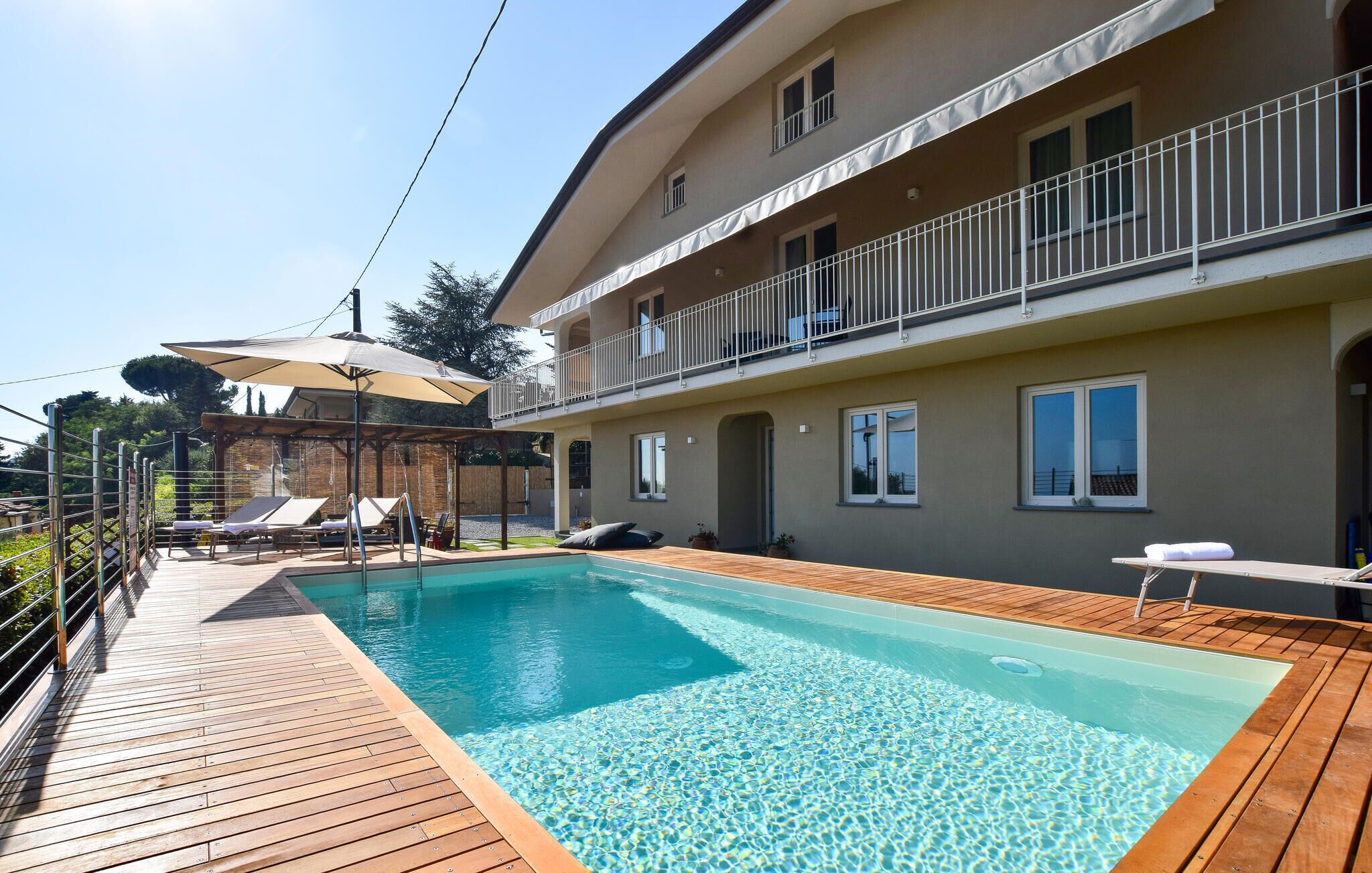 Ein Schwimmbad mit Holzterrasse, einer überdachten Sitzgruppe mit Sonnenschirmen und einem Gebäude mit Balkonen.