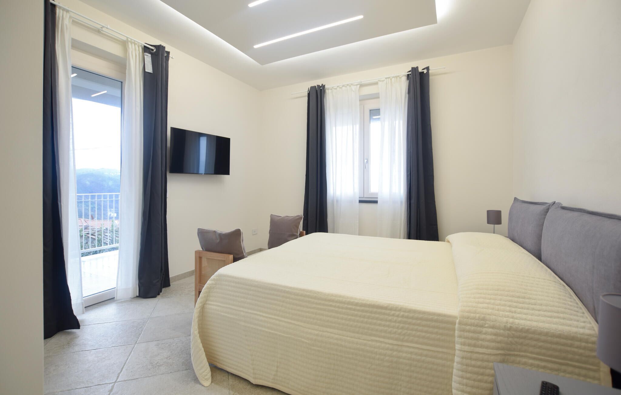 A hotel room with a large bed, a flat-screen TV mounted on the wall, a small bedside table, and a view of the outdoors through a balcony door.