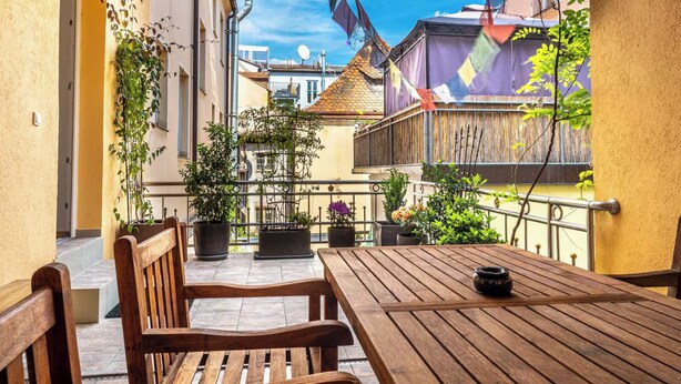 A wooden table and chairs on a balcony with potted plants and a view of buildings.
