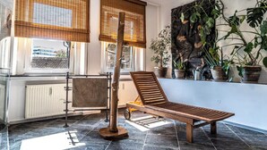 A sunroom with a wooden lounge chair, a radiator, a window with a bamboo blind, and potted plants.