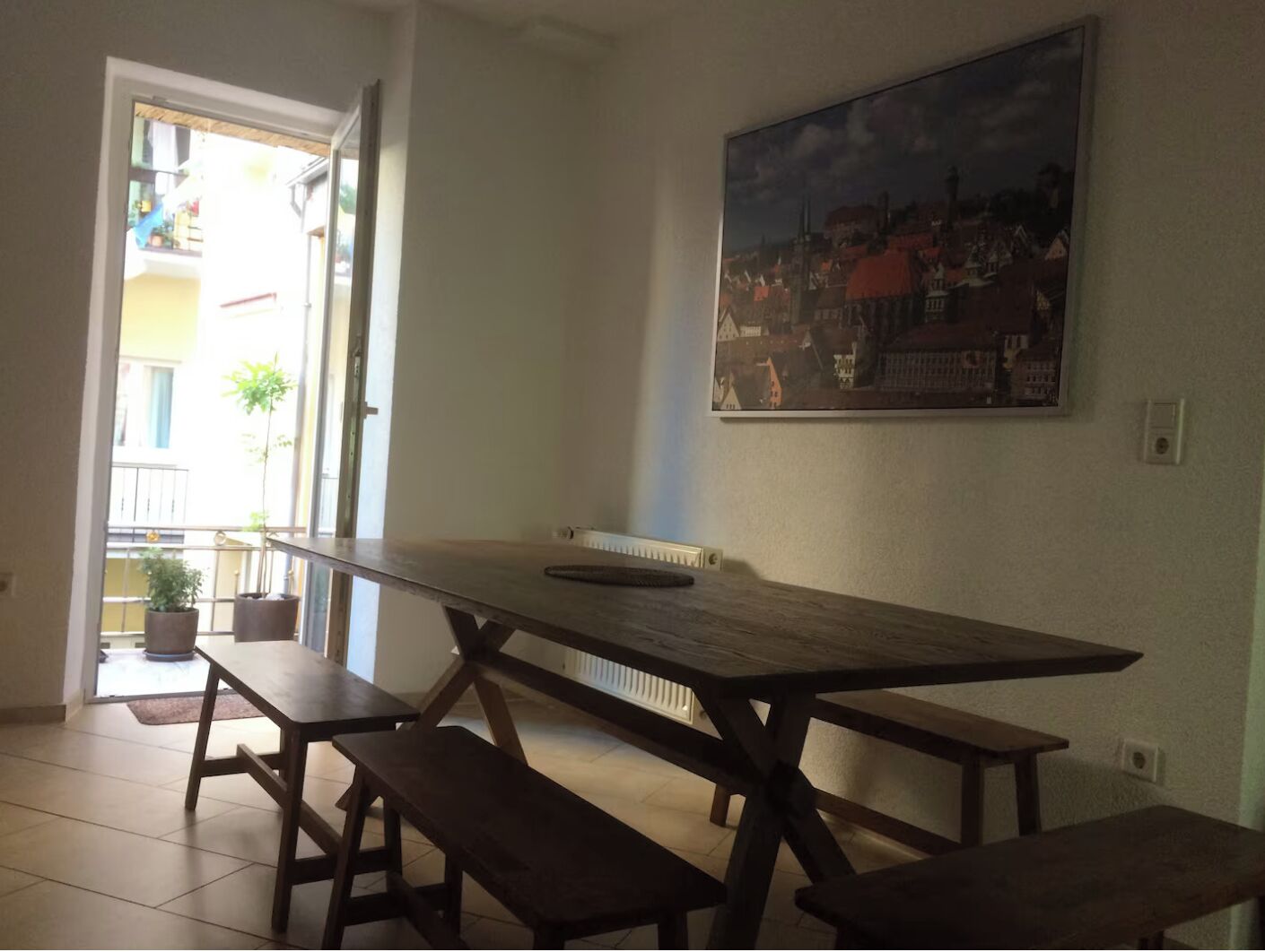 Un coin repas avec une grande table en bois et des bancs, un radiateur et un tableau représentant un paysage urbain accroché au mur.