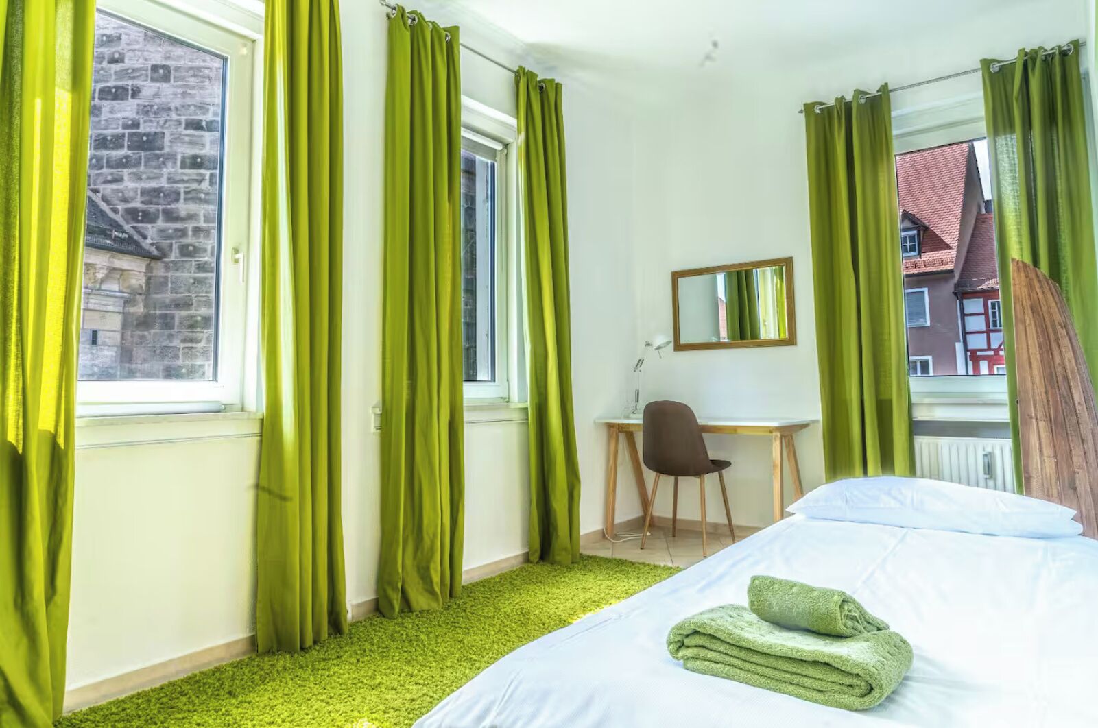 A hotel room with a bed, a desk, and a chair. There are curtains on the windows, and a mirror on the wall.
