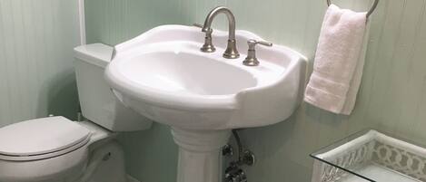 A bathroom with a pedestal sink, toilet, and a small glass shelf.
