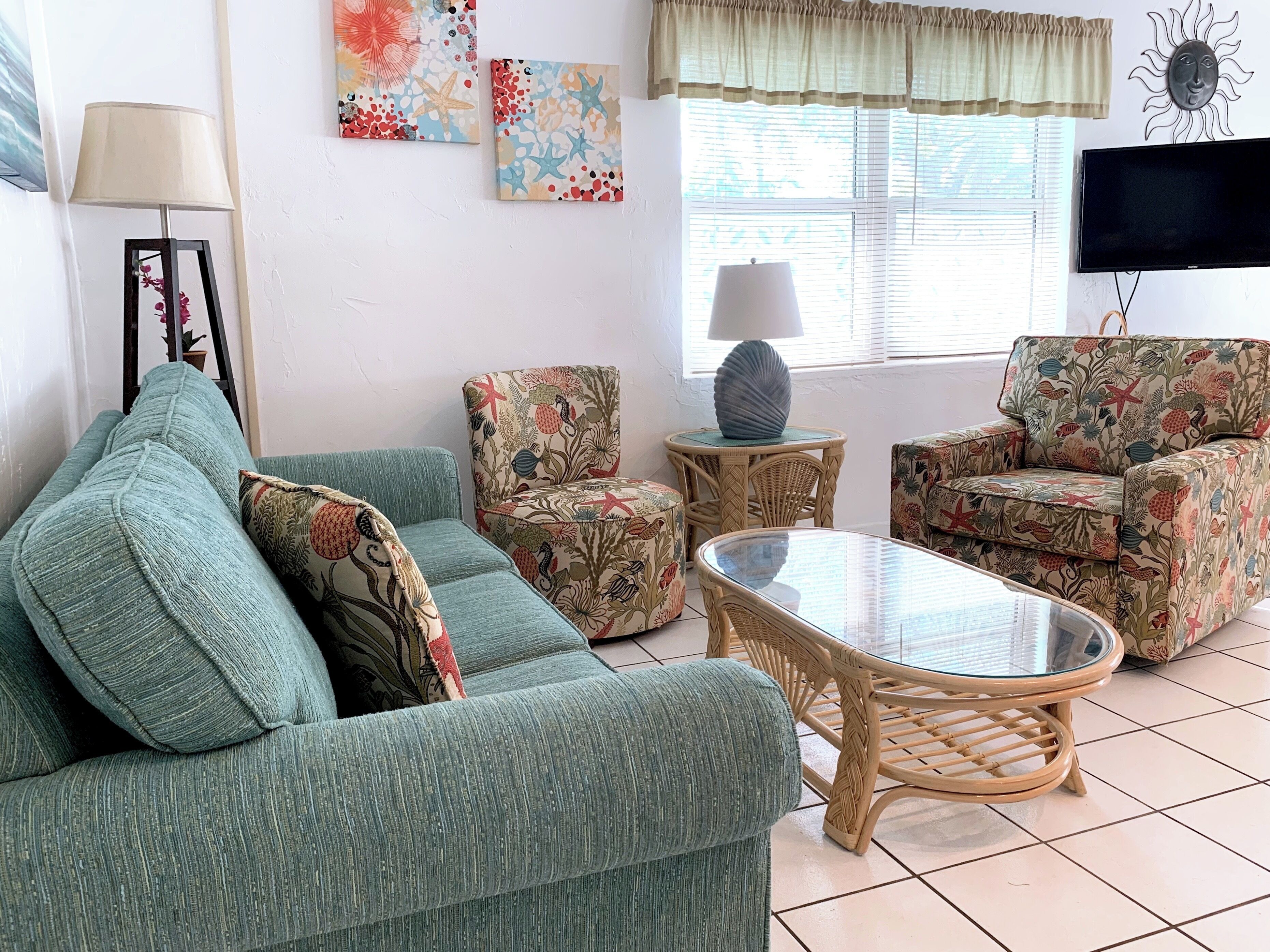 A living room with a green sofa, floral armchairs, a glass coffee table, and a TV.