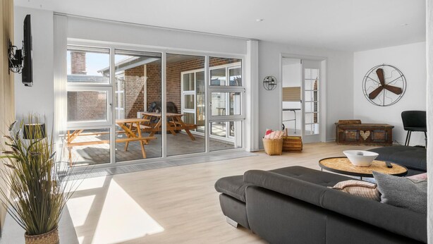 En moderne stue med en grå sofa, et spiseområde med træborde og en skydedør til en udendørs terrasse.