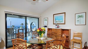 A dining area with a glass table surrounded by wooden chairs, a vase of flowers, and framed pictures on the wall. - Coastal Luxury @ Tyson Park | Ocean Views & Private Balcony (Oceanside)