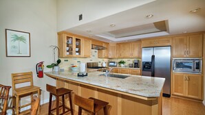 A modern kitchen with wooden cabinets, a stainless steel refrigerator, a microwave, and a large island with bar stools. - Coastal Luxury @ Tyson Park | Ocean Views & Private Balcony (Oceanside)