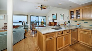 A spacious kitchen with wooden cabinets, a large island with a sink, and a view of the ocean through a sliding glass door. - Coastal Luxury @ Tyson Park | Ocean Views & Private Balcony (Oceanside)