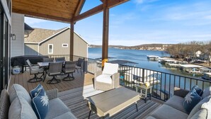 Eine überdachte Terrasse mit Gartenmöbeln, einem Grill und Blick auf einen See mit Stegen.