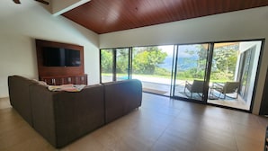A living room with a large flat-screen TV, a brown sofa, and a view of a pool and outdoor seating area through large glass doors. - Spacious 3 bed, 3 bath home with breathtaking Mountain and Valley views. (Platanillo)