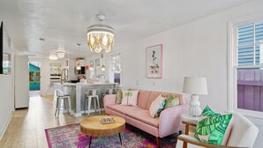 A cozy living room with a pink sofa, a round coffee table, and a vibrant rug. A kitchen area is visible in the background with modern appliances and bar stools.