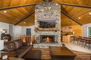 A cozy living room with a stone fireplace, wooden ceiling beams, a leather couch, and a dining area. - Charming Mountain Home with a View (Seven Devils)