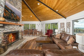 A spacious living room with a stone fireplace, leather furniture, wooden ceiling beams, and large windows offering a view of the outdoors. - Charming Mountain Home with a View (Seven Devils)