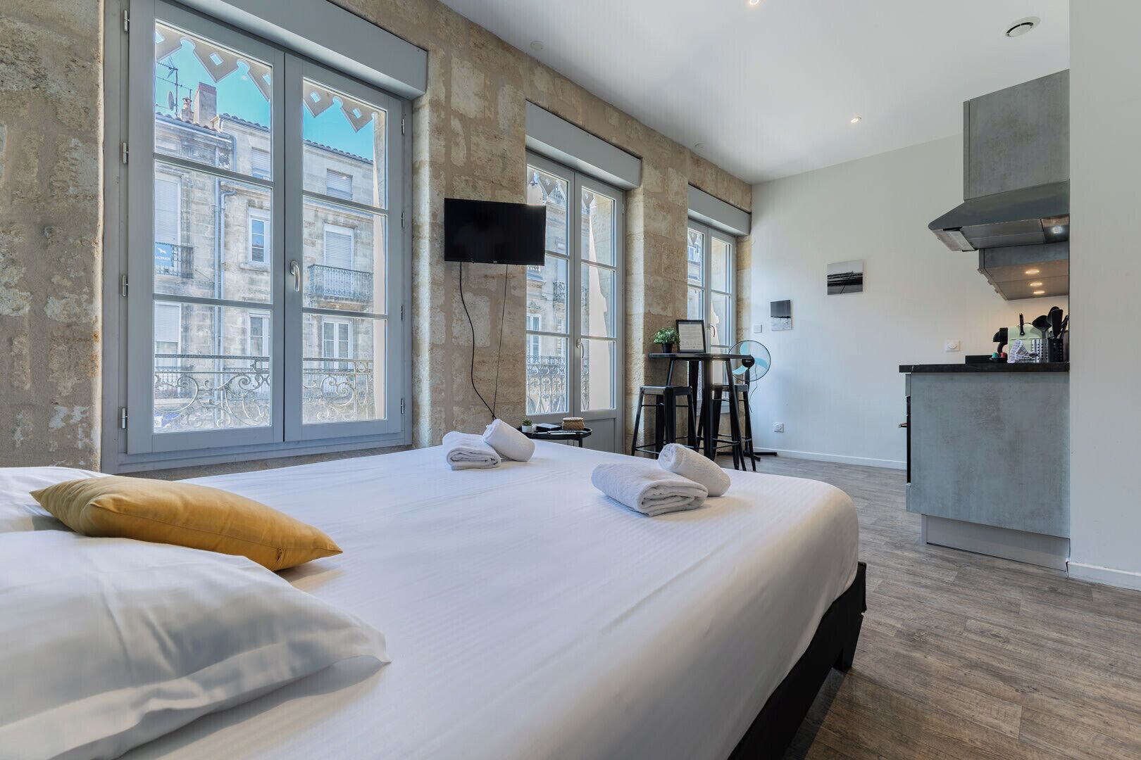 A modern hotel room with a large bed, a desk, a kitchenette, and large windows.
