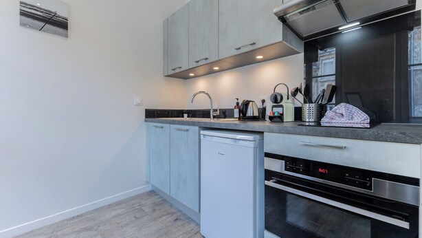 A modern kitchen with grey cabinets, a dishwasher, an oven, and a digital clock displaying 16:01. - Superb flat in beautiful Bordeaux stone for 2p (Bordeaux)