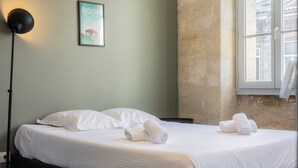 Un lit bien fait, avec du linge de lit blanc et des serviettes pliées, une lampe de sol noire et un tableau encadré au mur.