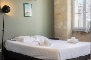 Een net opgemaakt bed met witte lakens en opgerolde handdoeken, een zwarte vloerlamp en een ingelijst fotobehang aan de muur.