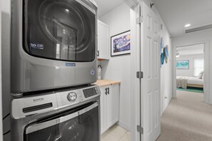 Kompakti pyykinhuone, jossa on Maytag-merkkinen pesukone ja -kuivausrumpu, puinen työtaso sekä ovi, joka johtaa makuuhuoneeseen, jossa on sänky ja seinällä taulu.