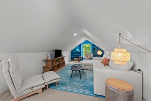 A cozy attic bedroom with a large window, a sofa, a chair, a small table, and a TV. - Walk on Water: Soundside, Community Pool and Clubhouse Access (Kill Devil Hills)