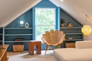 Een gezellig slaapkamer op zolder met een schuine plafond, een groot raam, een rieten stoel, een klein tafeltje en een bedlampje.